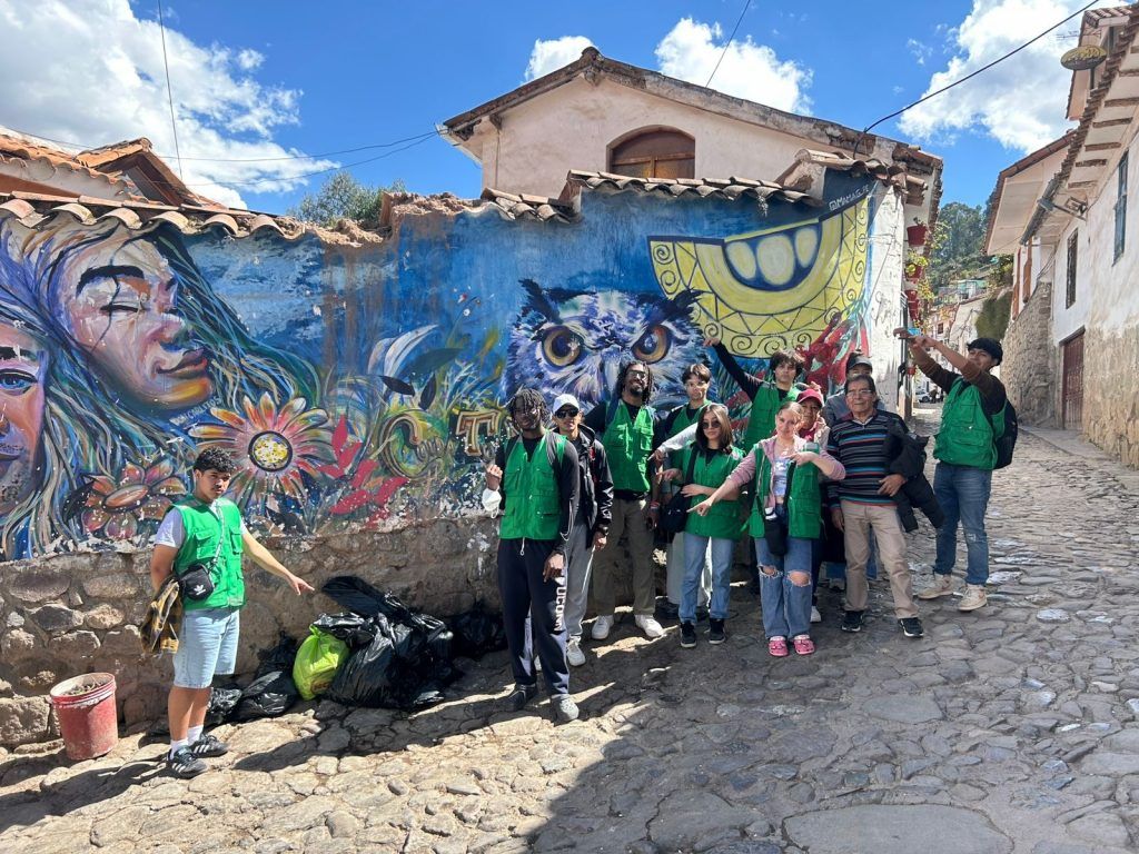 Micheal Orejuela 28 (CLAS) in Cusco, Peru, in summer 2025. Student group standing on a cobblestone street in front of a multicolored mural with peoples' faces, an owl, grass and other shapes.