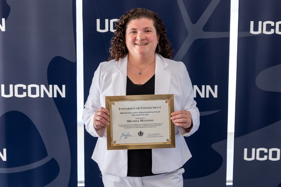 Melissa Manning is holding her NACADA Region 1 Advising Excellence Award in front of a blue and white UCONN backdrop.